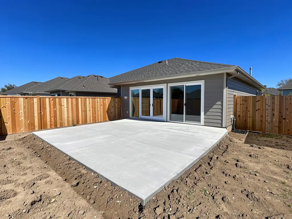 Before: a plain concrete patio behind a new home, with bare soil and a cedar fence